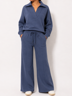 A person wearing a matching two-piece loungewear set in slate blue, featuring a long-sleeve collared top and wide-leg drawstring pants, paired with off-white slip-on shoes against a plain light background.